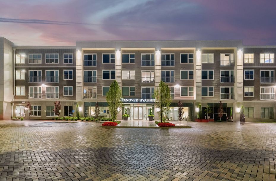 Hanover Company, Hanover Hyannis Apartment building entrance, "Hanover Hyannis," with illuminated façade and a wide brick-paved plaza at dusk.