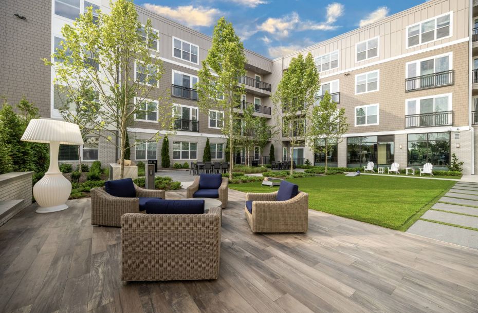 Hanover Company, Hanover at the Pinehills Modern apartment courtyard with wicker chairs, green lawn, trees, and a large white lamp under a blue sky.