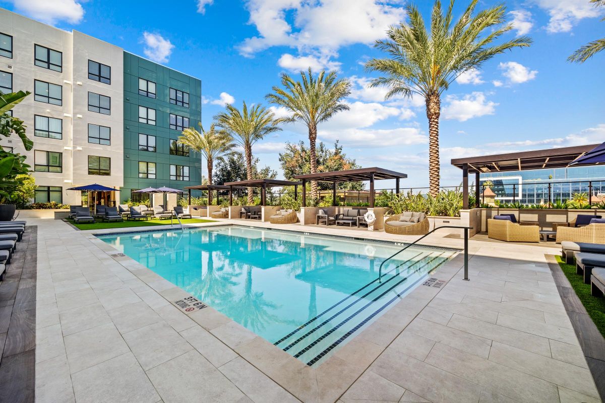 Outdoor pool with lounge chairs, cabanas, and palm trees near a modern building under a blue sky with clouds.