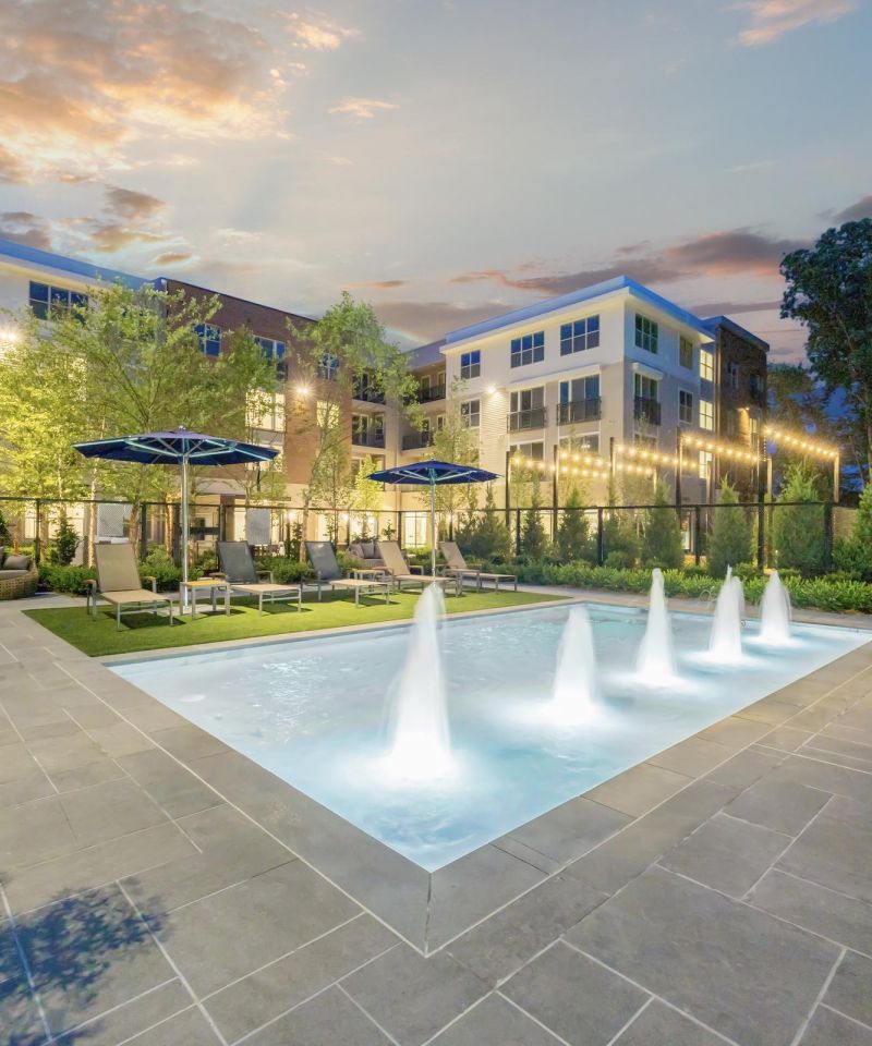 Modern apartment complex with a lit pool, water jets, lounge chairs, and evening sky.