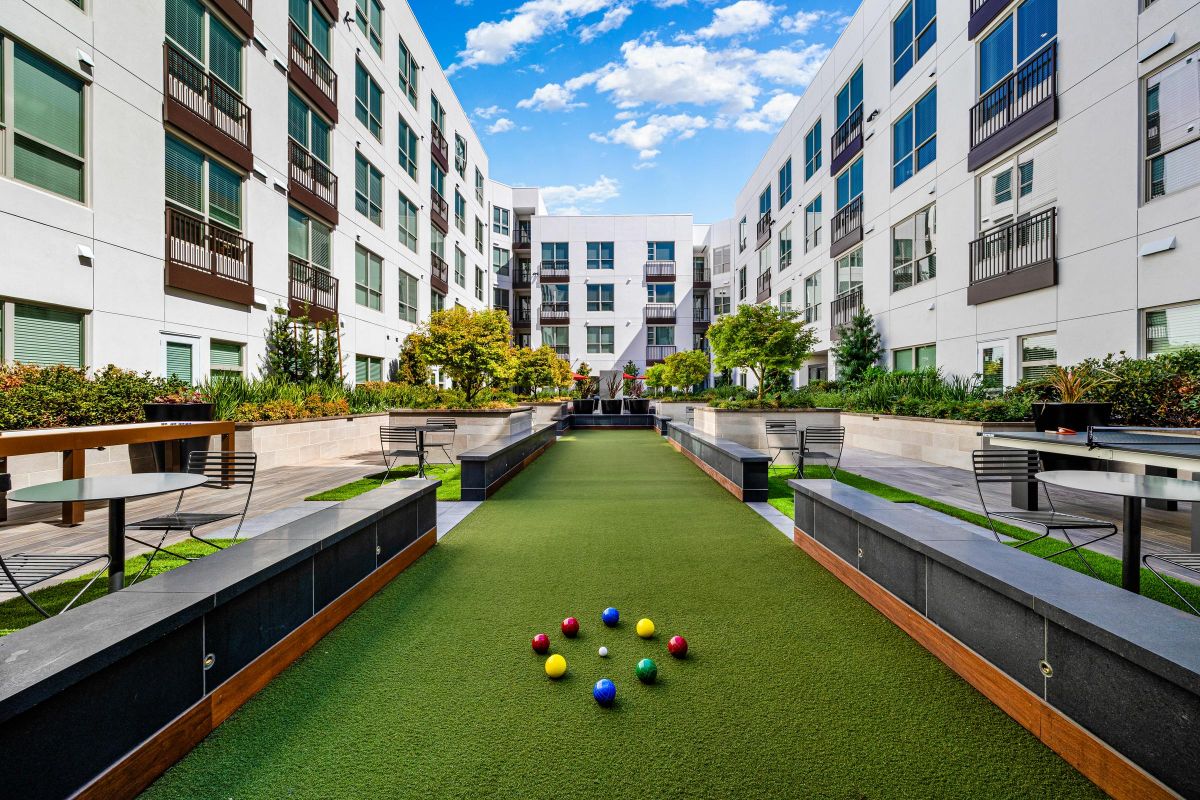 Courtyard with bocce ball setup, surrounded by modern apartments, outdoor seating, and greenery under a blue sky.