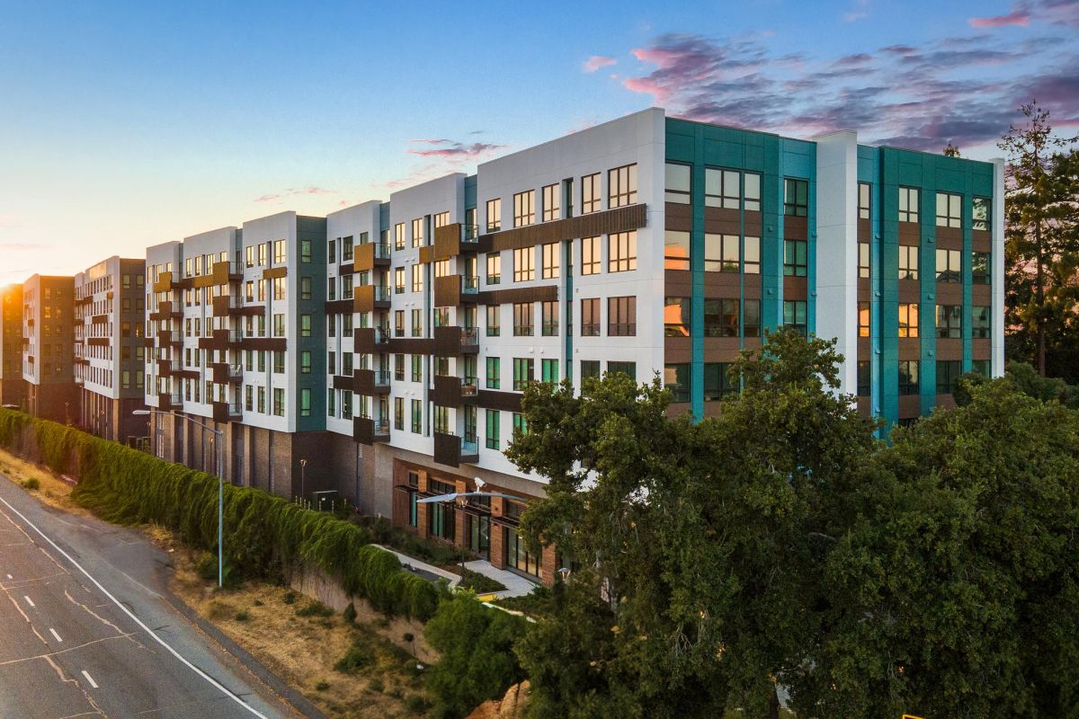 Modern apartment complex with large windows and balconies at sunset, adjacent to a road and surrounded by trees.