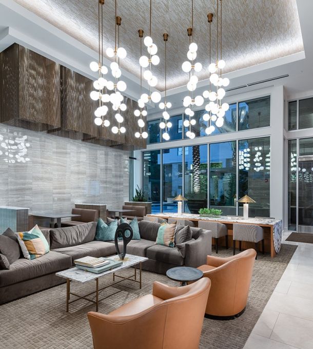 Hanover Company Modern lobby with gray sofas, tan armchairs, bright spherical chandeliers, and large windows reflecting city lights.