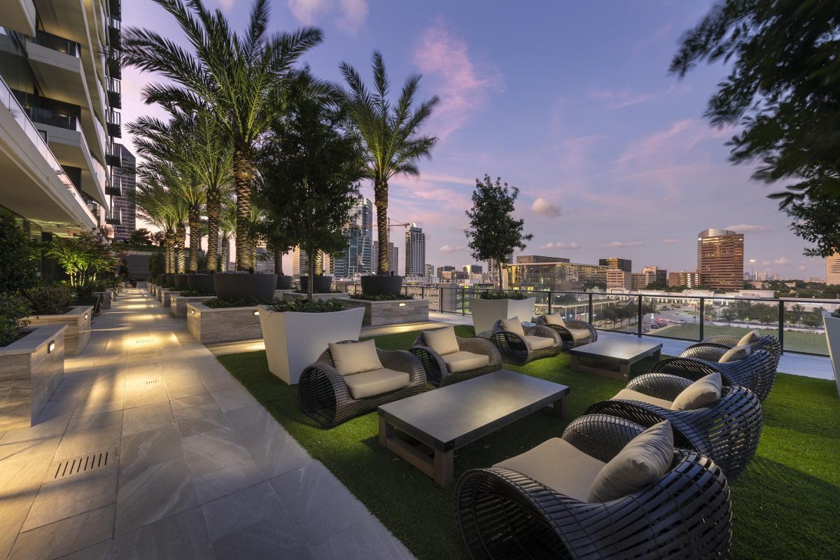 Rooftop patio with wicker chairs, tables, palm trees, and city skyline view at dusk.