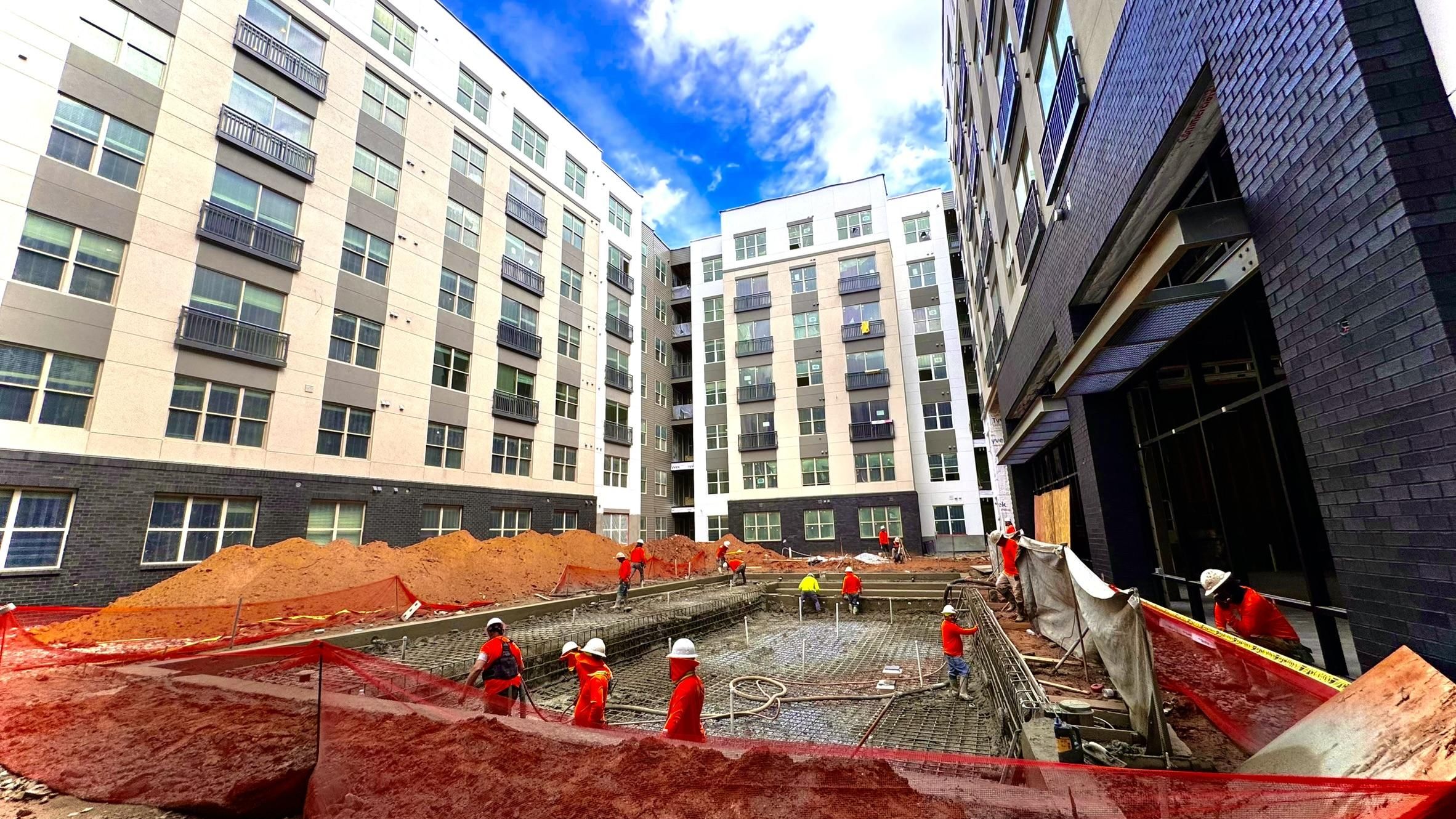 Construction workers in hard hats work on a building site surrounded by tall apartment buildings.