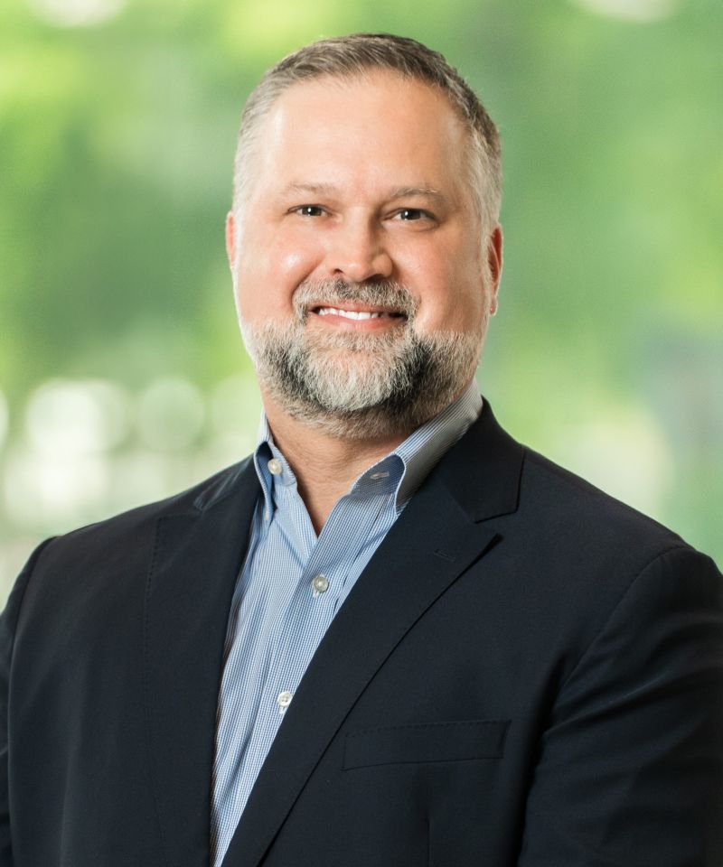 Smiling man with gray beard in a suit jacket and collared shirt standing in front of a blurred green background.