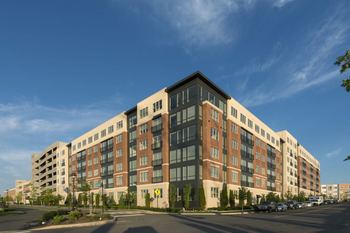 Modern multi-story apartment building on a sunny day, with cars parked along the street and blue sky above.