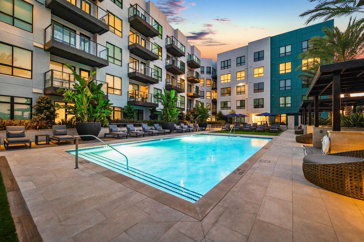 Modern apartment complex with a lit swimming pool, lounge chairs, and sunset sky in the background.