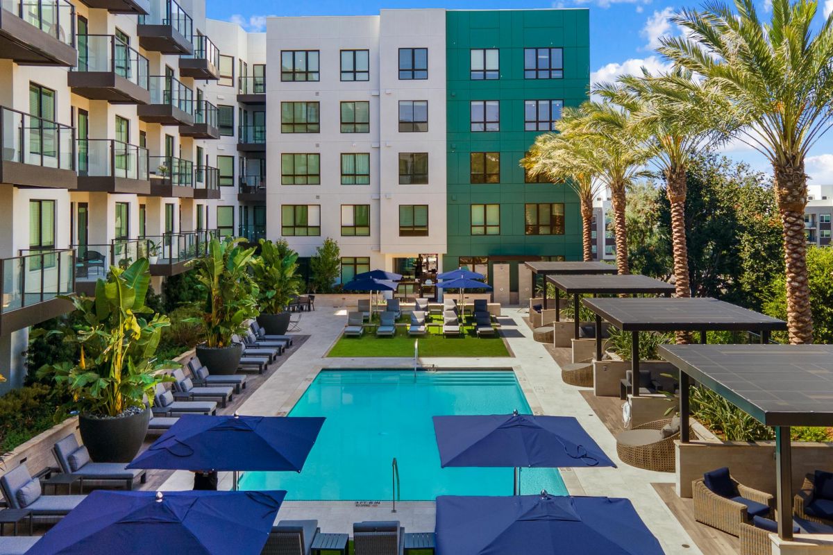 Modern apartment complex with a central pool, sun loungers, umbrellas, and palm trees under a blue sky.
