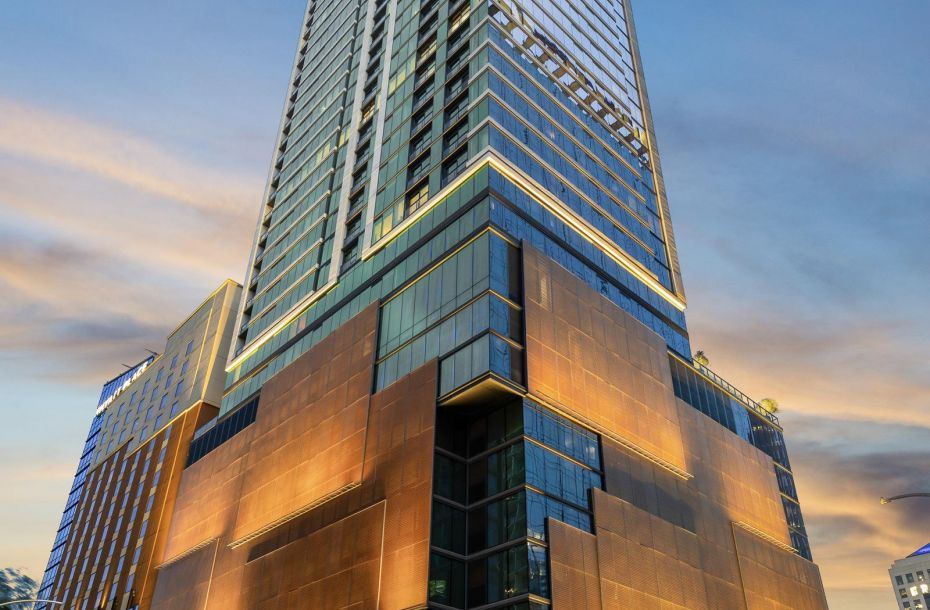 A tall, modern skyscraper with a glowing exterior stands against a twilight sky, surrounded by city lights and cars.