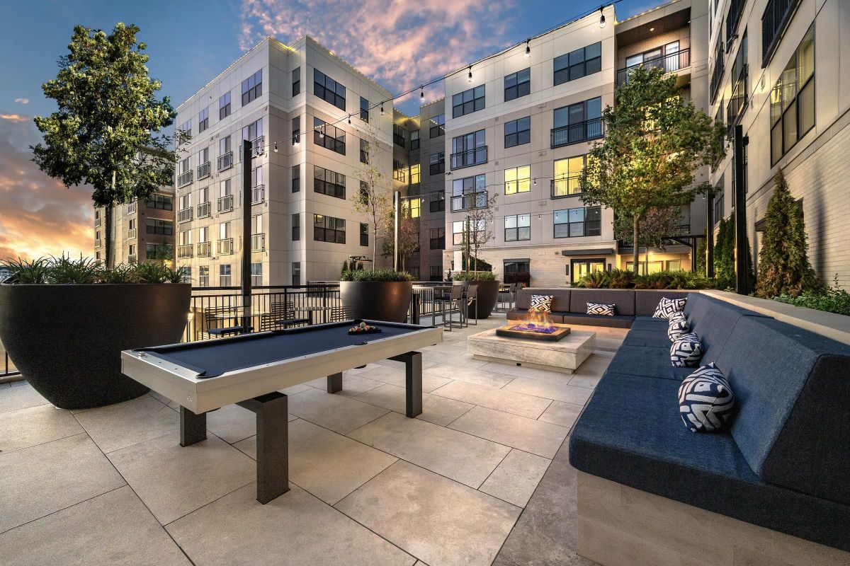 Modern outdoor patio with a pool table, fire pit, and seating, surrounded by tall buildings at sunset.