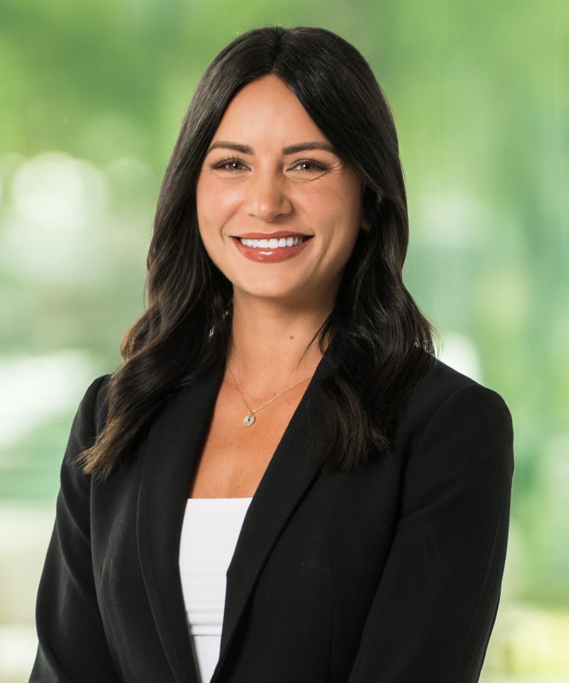 Chelsie Ruggieri, in a beige blazer, smiles indoors with a blurred green background.