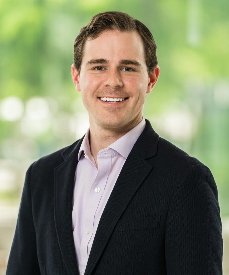 Smiling man in a suit jacket and light purple shirt, standing in front of a blurred green background.