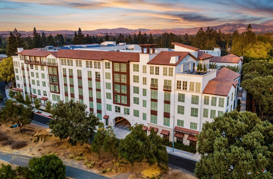 Hanover Company, Hanover Walnut Creek Large multi-story building surrounded by trees, against a backdrop of rolling hills and a colorful sunset sky.