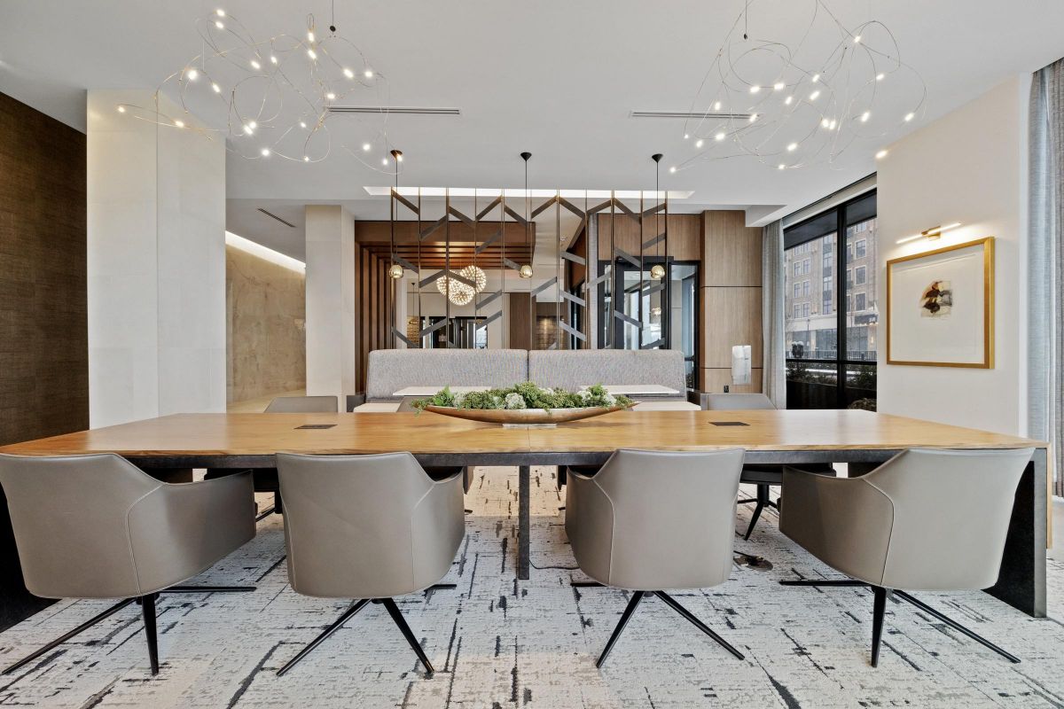 Modern conference room with a long wooden table, beige chairs, and artistic lighting fixtures. Large windows on the right.