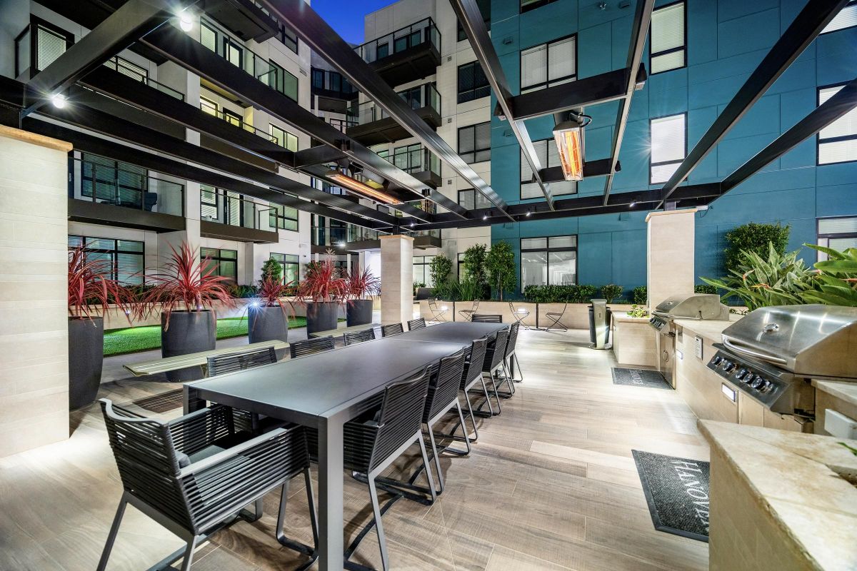 Modern outdoor patio with a long dining table, chairs, grills, and planters; surrounded by a building facade.
