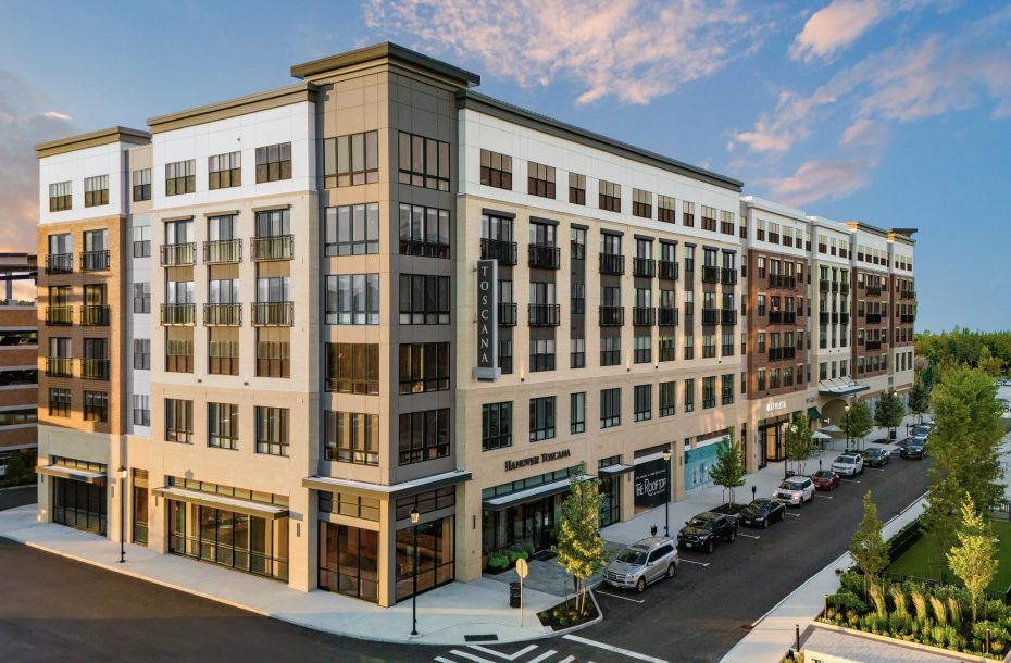 Modern multi-story building with large windows, lined by parked cars and trees under a blue sky.