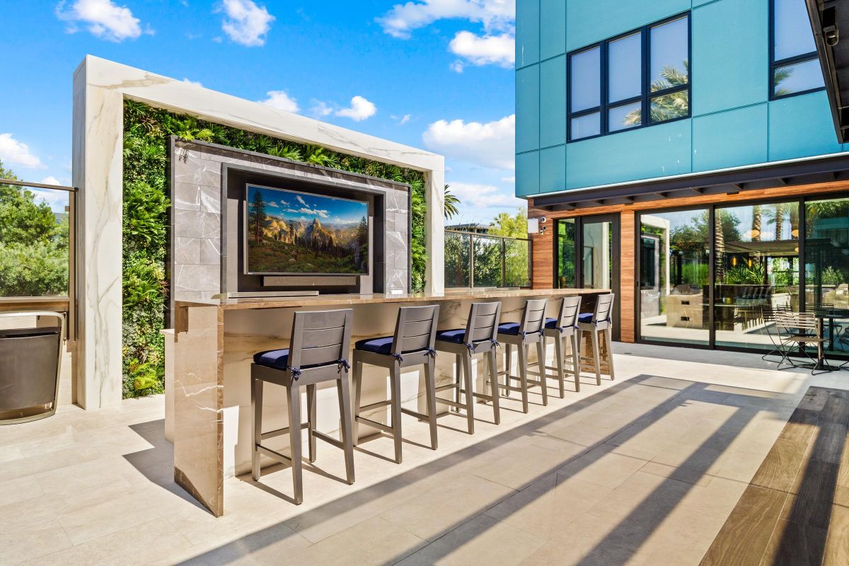 Outdoor patio with a bar counter, high chairs, wall-mounted TV, and lush greenery, under a clear blue sky.