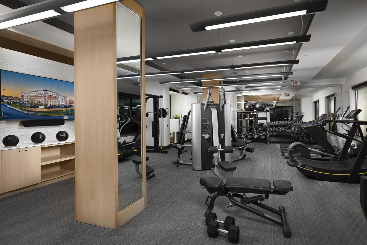 Modern gym with exercise machines, weights, benches, and large mirrors. Bright lighting and a wall-mounted TV.