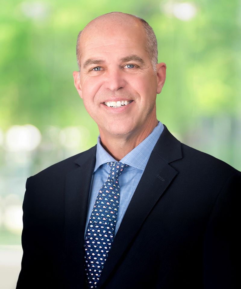 Smiling man in a suit and tie standing in front of a blurred green outdoor background.