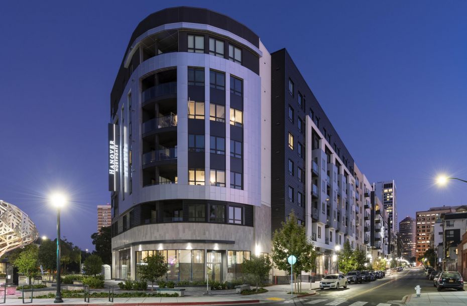 Modern apartment building on a city street corner at dusk, with streetlights and trees.