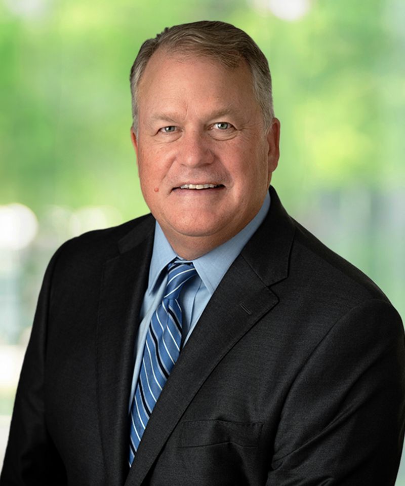 Smiling man in a suit and blue striped tie poses in front of a blurred green outdoor background.