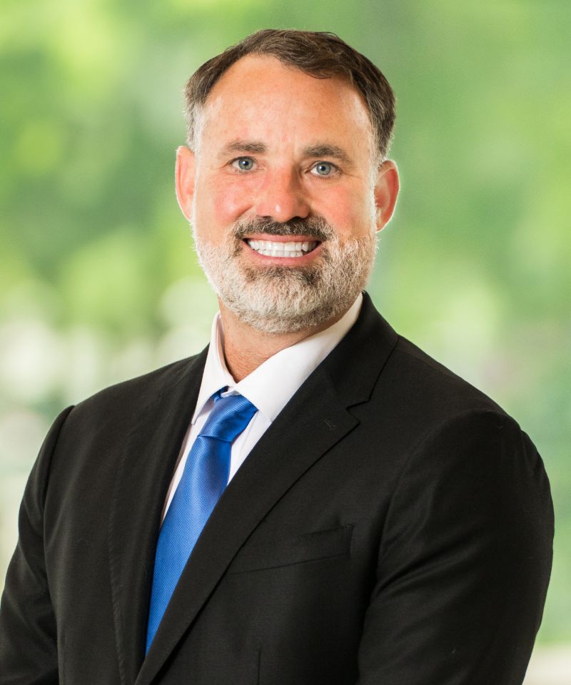 Smiling man in a suit and blue tie, standing in front of a blurred green outdoor background.