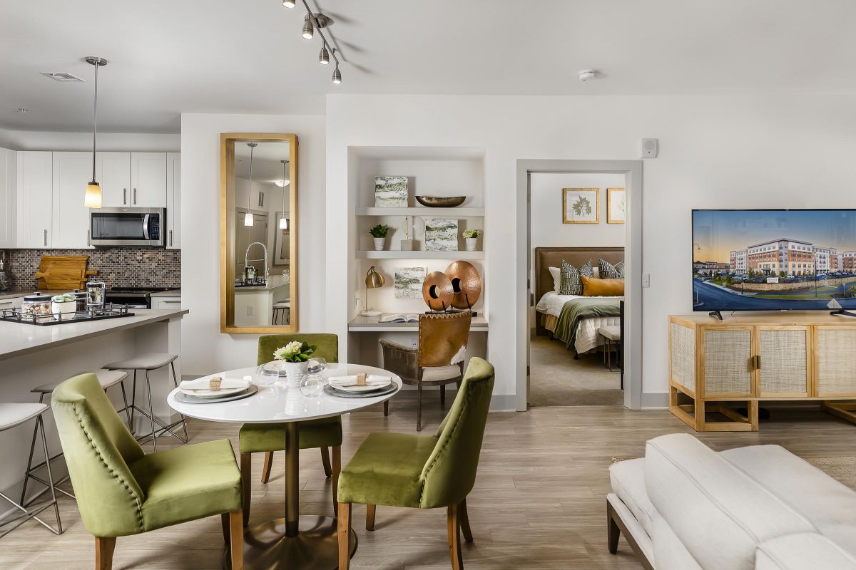 Modern open-plan apartment with kitchen, dining area, workspace, and bedroom; green chairs and neutral decor.