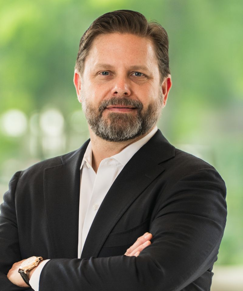 Man with brown hair and beard in a suit, standing with arms crossed, blurred green background.