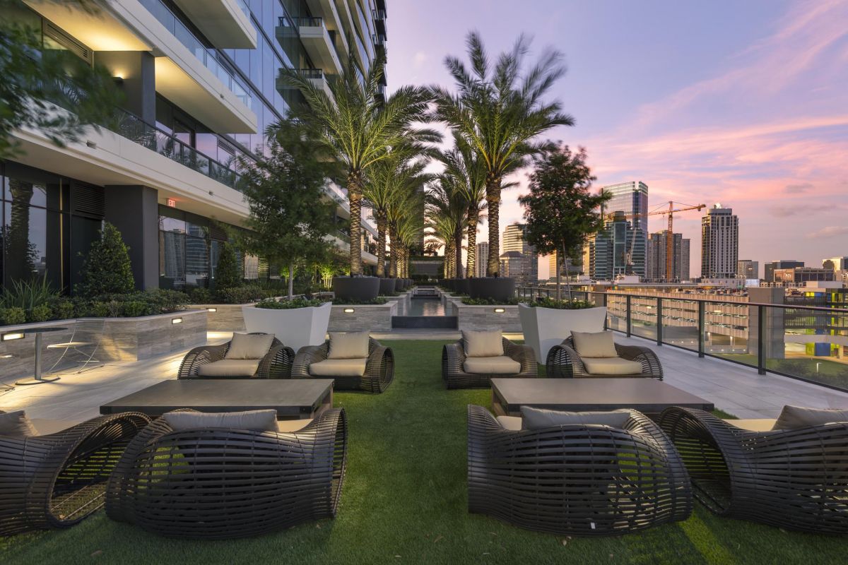 A modern rooftop terrace with wicker chairs, palm trees, and city skyline at sunset.