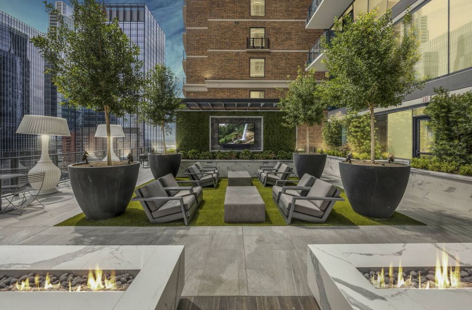 Hanover Company, Hanover Republic Square Modern rooftop patio with seating, planters, fire pits, and a large screen, surrounded by city buildings at dusk.