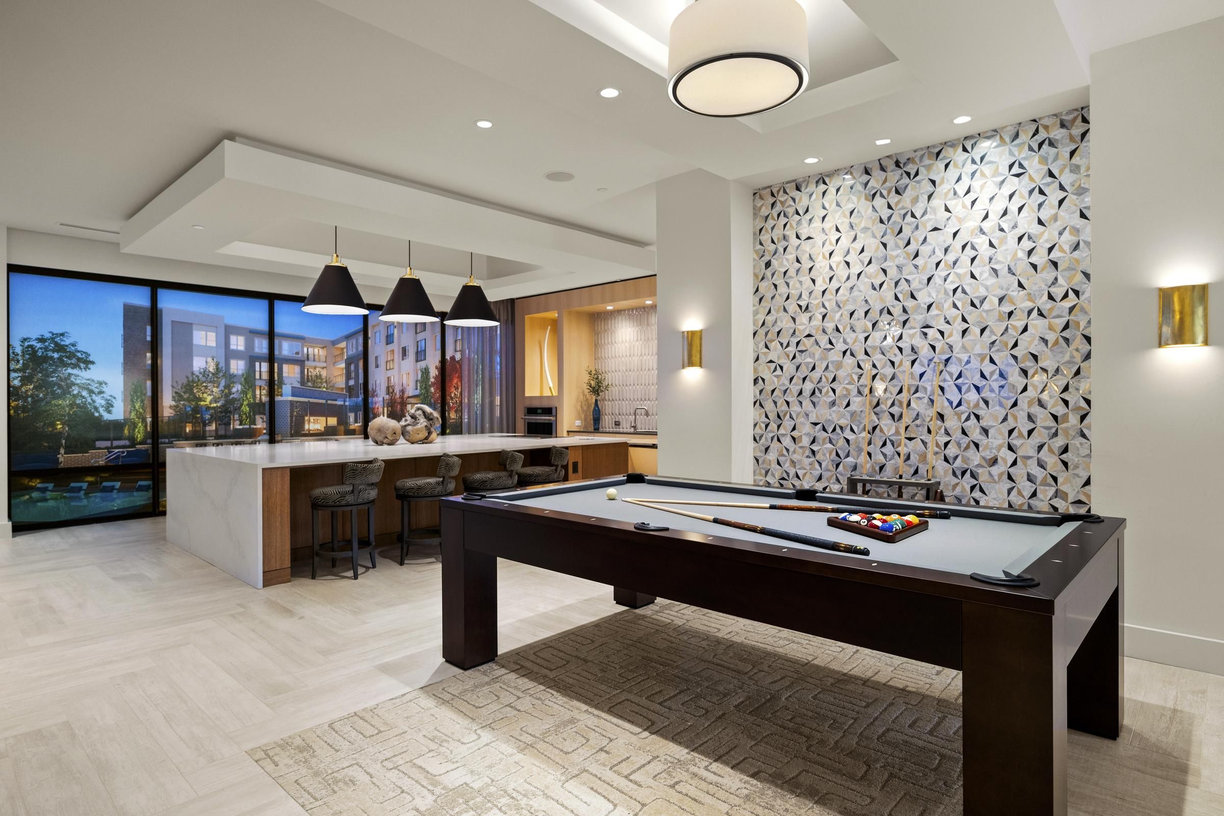Modern game room with a pool table, bar area, geometric tiled wall, and large windows overlooking a building at Hanover Colony Place.