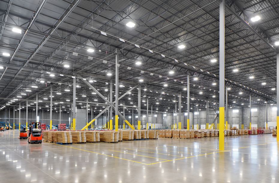 Large, brightly lit warehouse with high ceilings, forklifts, and stacks of pallets and boxes.