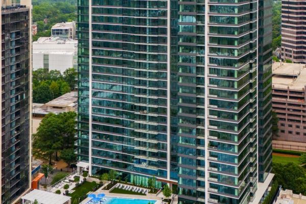 Tall glass apartment building with a pool and trees at its base, surrounded by other high-rise buildings.