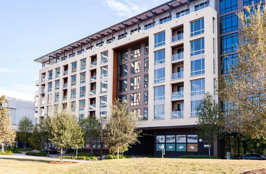 Modern mid-rise apartment building with balconies, large windows, and trees in the foreground.